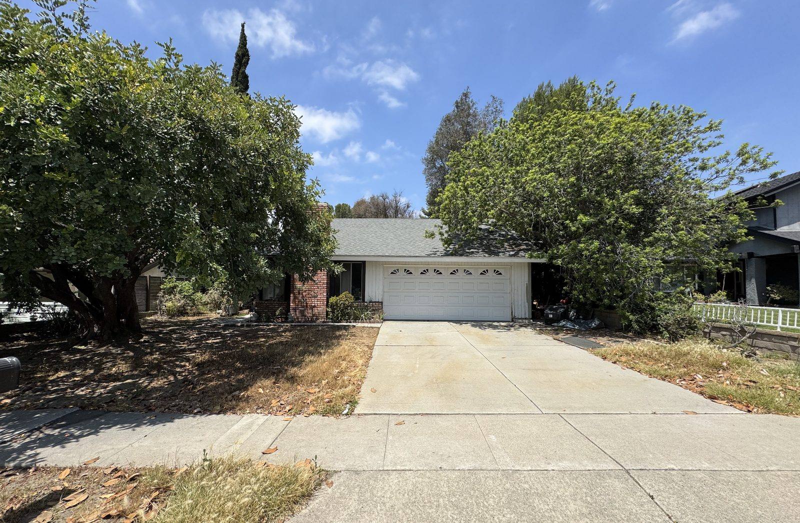 The house from the street before work: overgrown trees hiding the front, dead lawn, a tired white garage door.