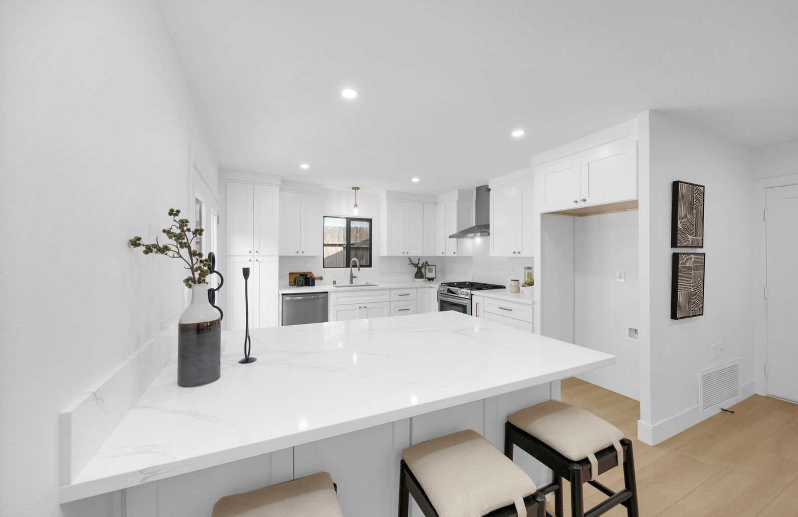 The kitchen after: a wall removed, an island with a stone worktop and stools, white shaker cabinets, stainless appliances and a range hood.
