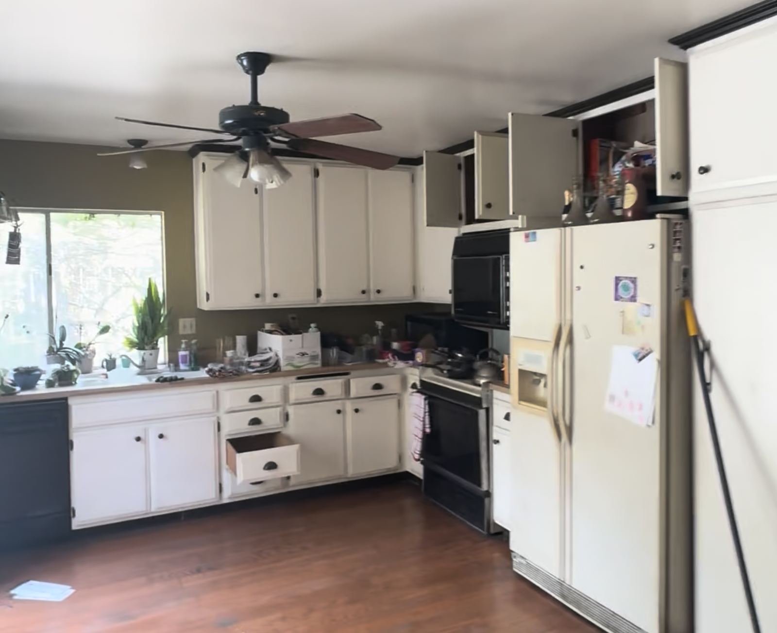 The kitchen before: dated cabinets, laminate worktops, a ceiling fan, mismatched black appliances and an ageing fridge, counters covered in belongings.