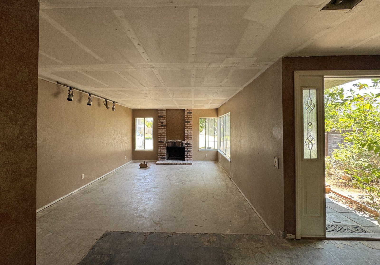 The living room stripped back: bare concrete floor, brown stucco walls, an original brick fireplace and exposed drywall seams across the ceiling.